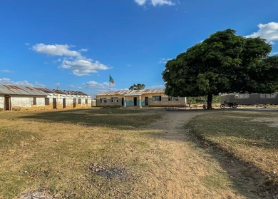 Rural school campus with several simple buildings, a flagpole with flag, a large tree, and dry grassy ground under a blue sky