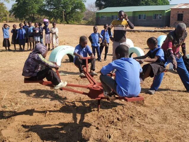 Kinder spielen auf einem staubigen Schulhof an einem Karussell und bemalten Reifen, im Hintergrund stehen weitere Kinder