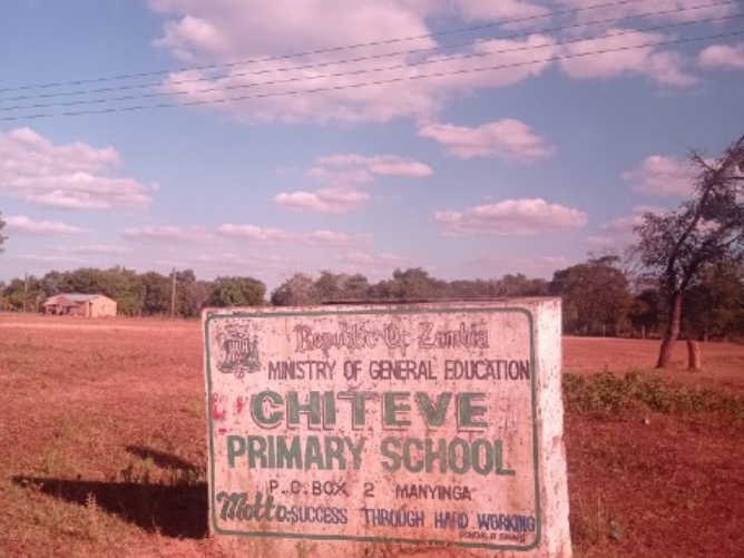 Ein verwittertes Schild der Chiteve Primary School in Sambia steht auf trockenem, rötlichem Boden unter blauem Himmel