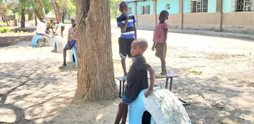 Mehrere Kinder spielen auf einem staubigen Platz mit bemalten Reifen und Bänken, im Hintergrund ein Gebäude und Bäume